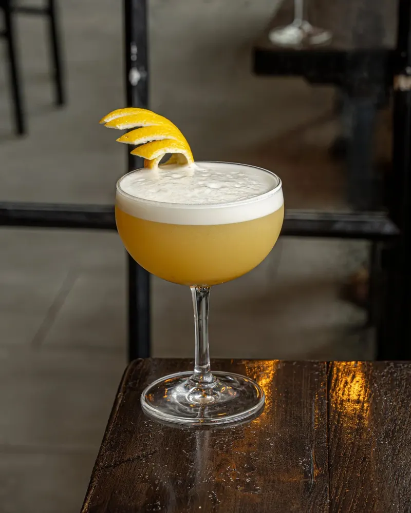 A yellow, foamy cocktail with a lemon twist on a wooden bar