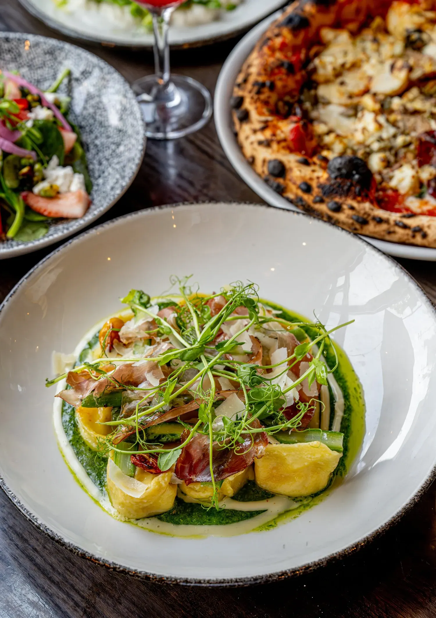 Overhead table of wood-fired pizza, filled pasta with prosciutto and greens, a salad and a glass of wine